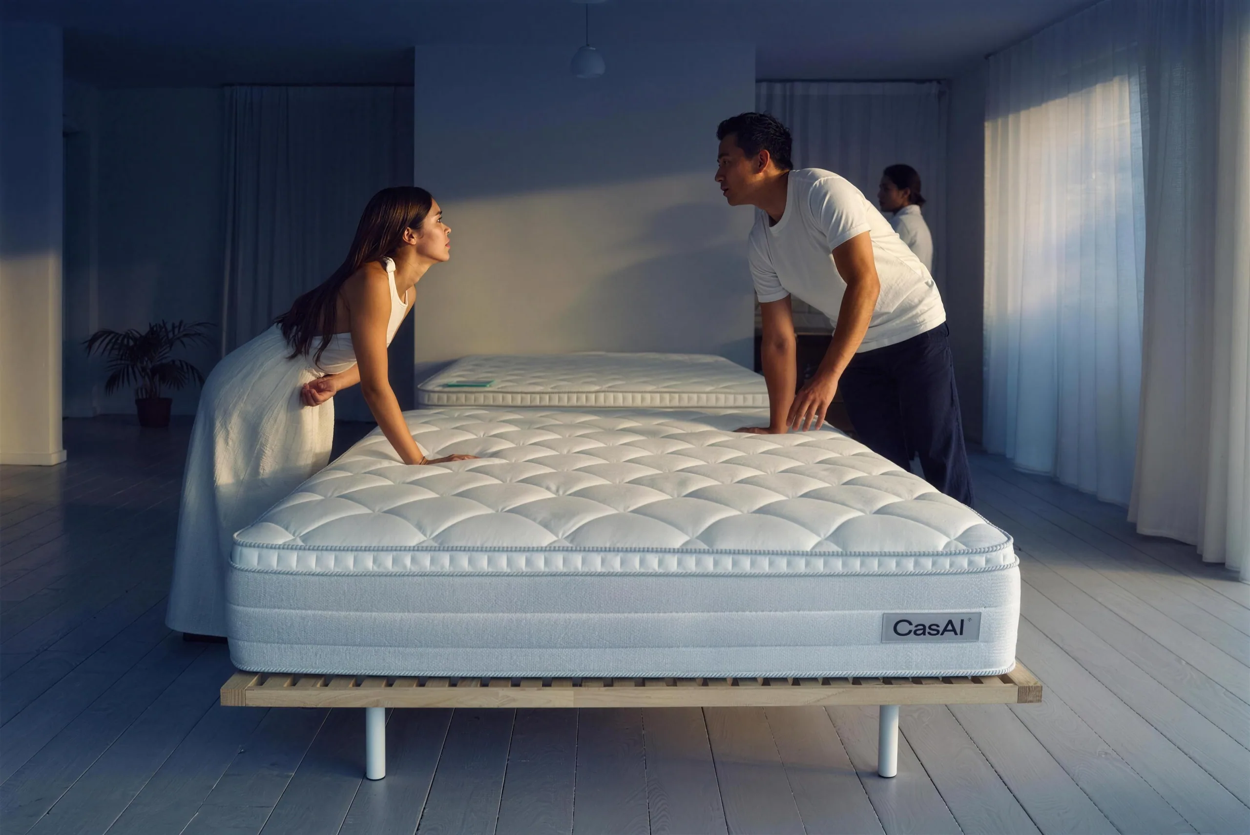 A couple in a bright mattress showroom, each pressing a hand into a different mattress, a salesperson out of focus behind them. (illustration)