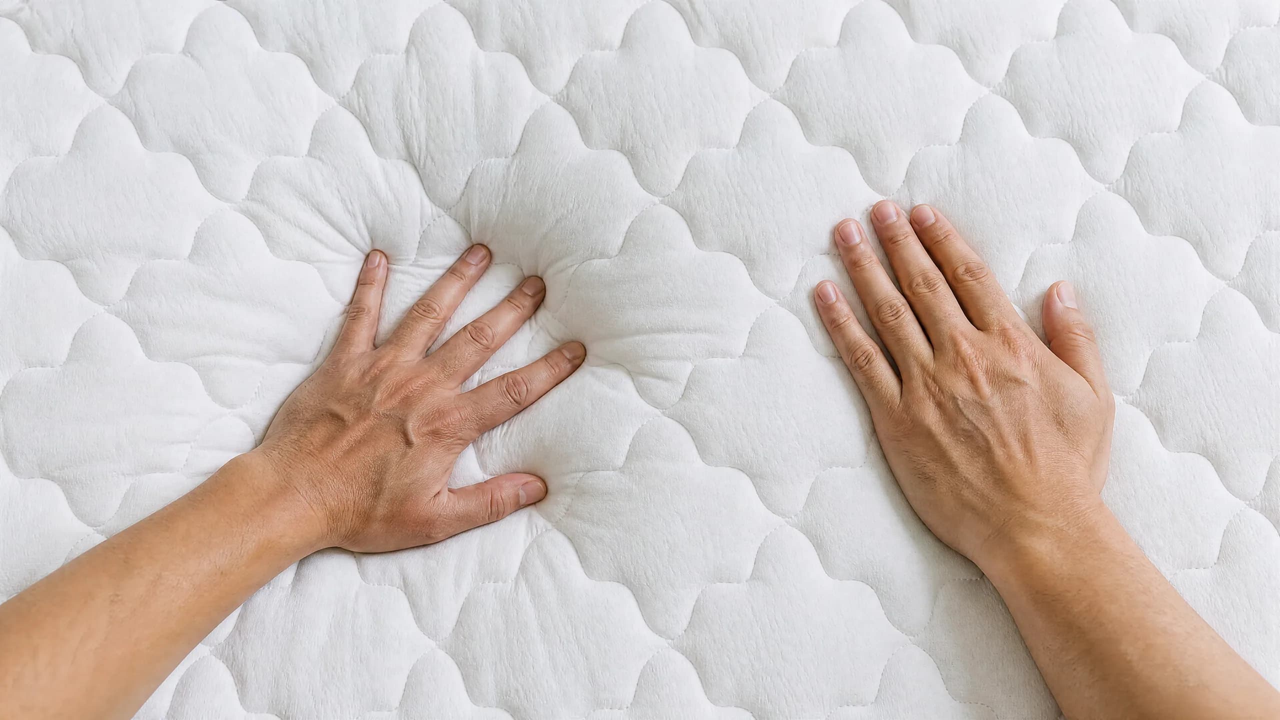 Two hands on one mattress from above, one pressing hard and one resting lightly, the quilt deforming differently under each. (illustration)