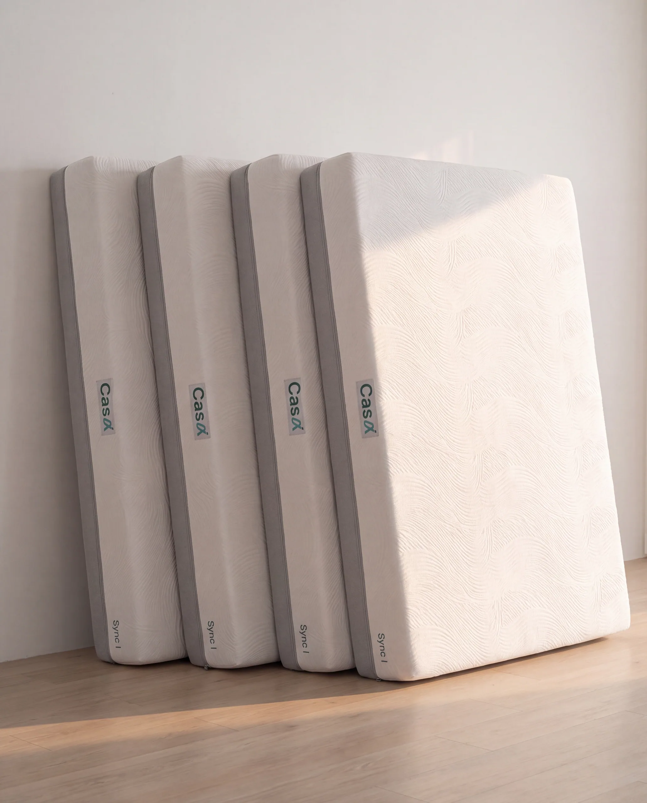 Four Sync mattresses leaning in a stagger against a white showroom wall in morning light.